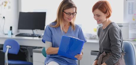 Clinician sat down with a patient. Both of them are looking at a blue clip board that the clinician is holding. 