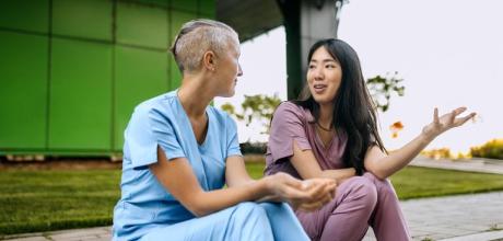 Two nurses in scrubs chat outdoors. One with short hair, one gesturing whilst speaking.