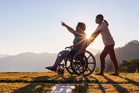 Caregiver pushes a person in a wheelchair pointing at a bright sunset over hills.