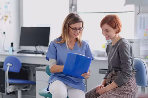 Clinician sat down with a patient. Both of them are looking at a blue clip board that the clinician is holding. 