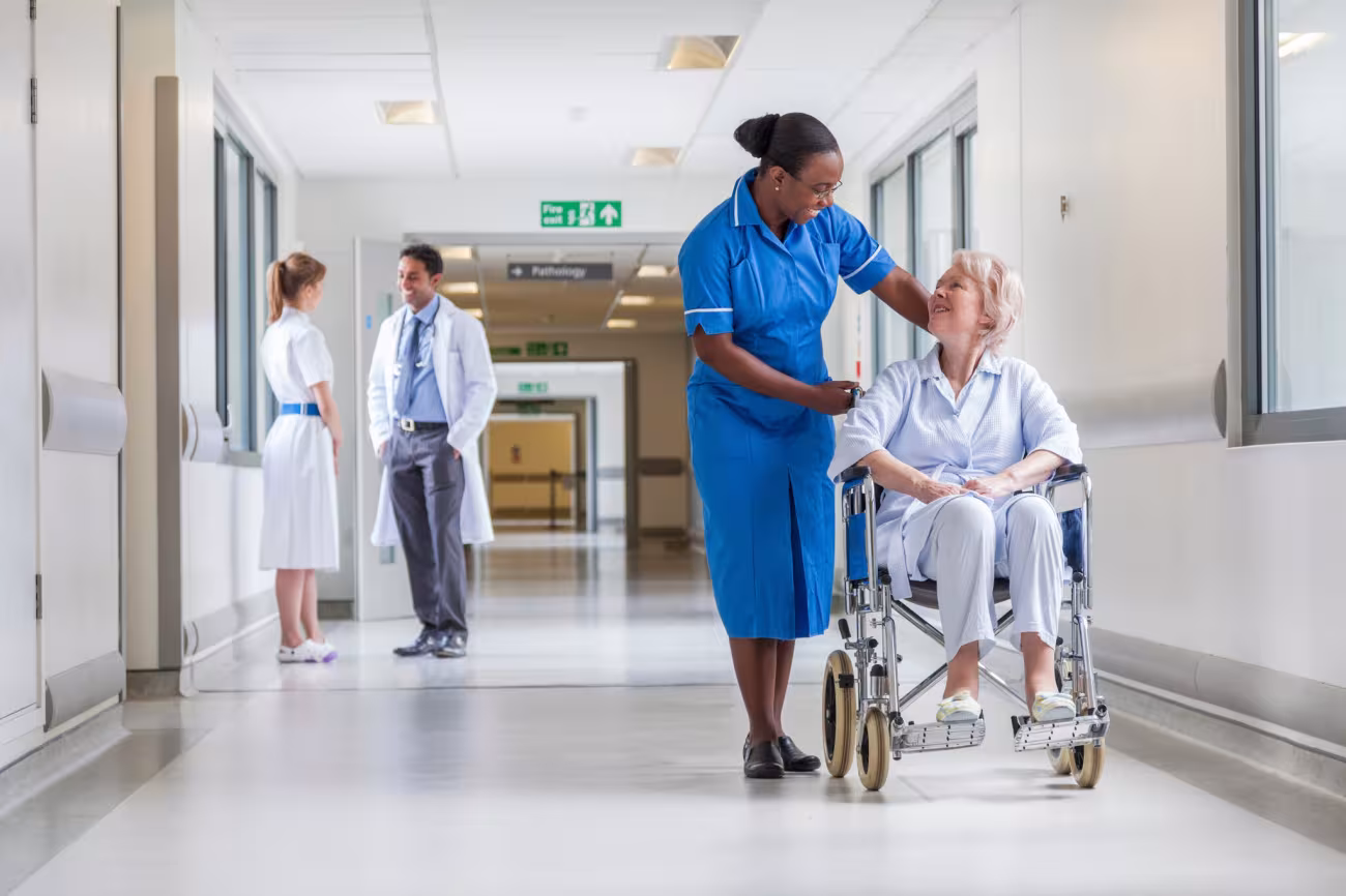 Photograph: Nurse assists an elderly woman in a wheelchair along a bright hospital corridor.