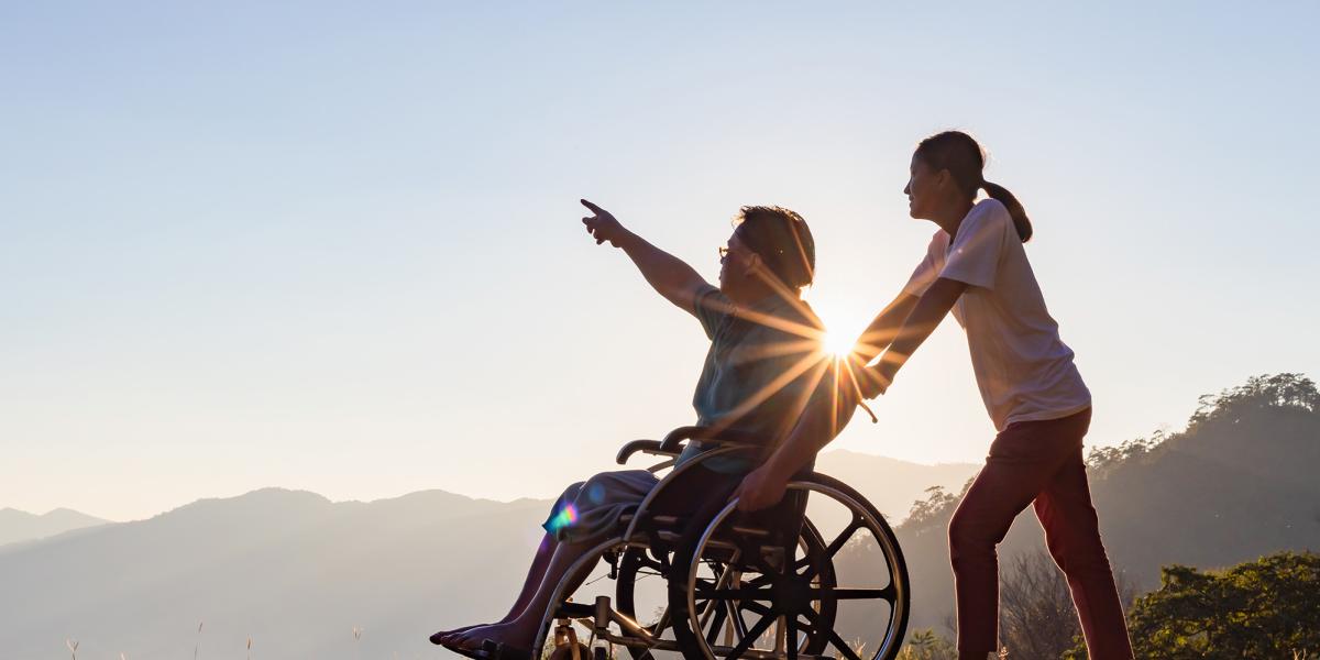 Caregiver pushes a person in a wheelchair pointing at a bright sunset over hills.