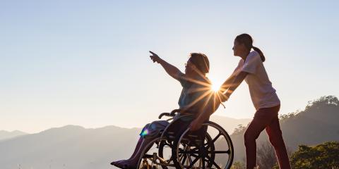 Caregiver pushes a person in a wheelchair pointing at a bright sunset over hills.